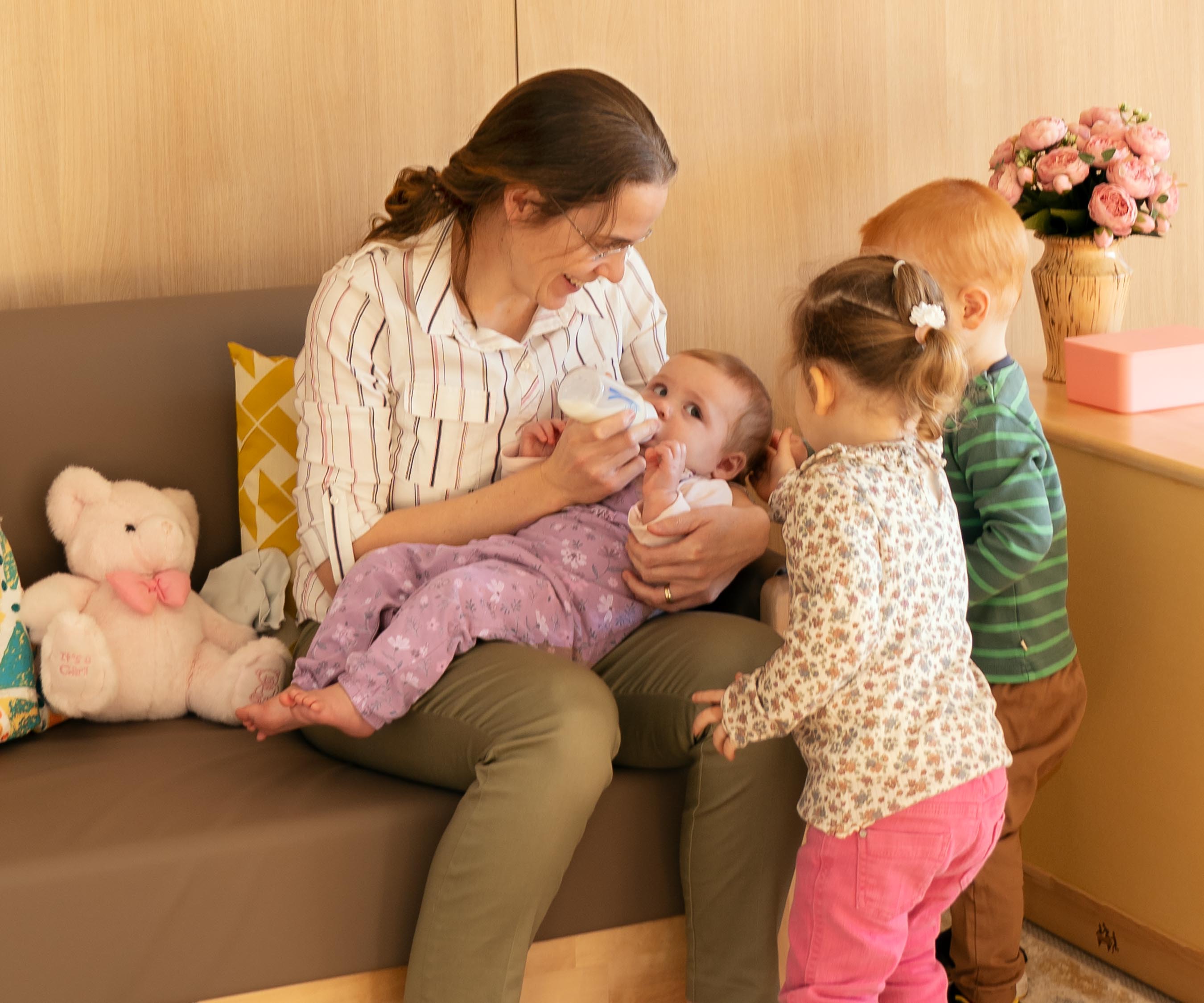 A nursery teacher seated on a natural-coloured sofa and feeding a baby a bottle