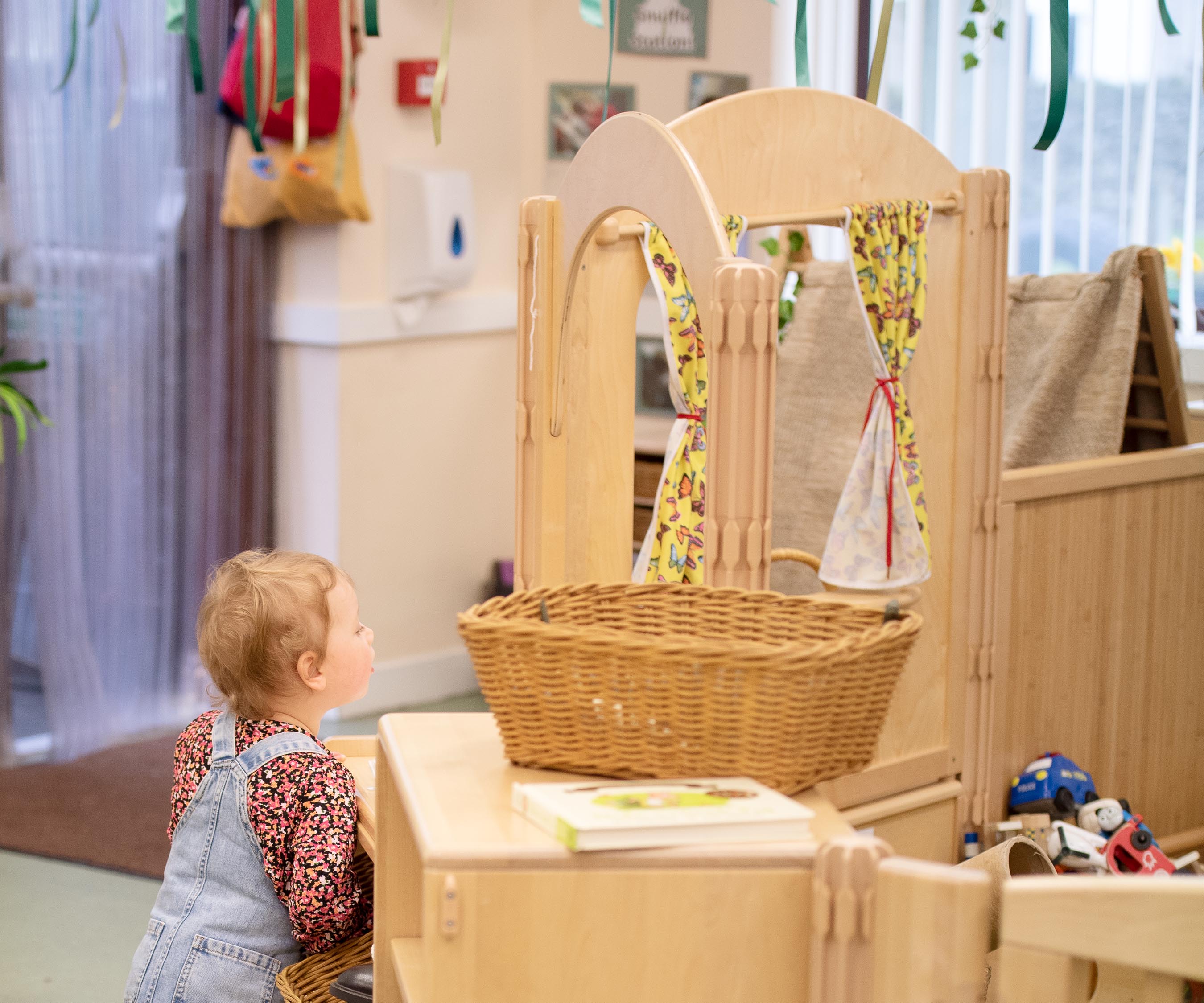 toddler girl wearing overalls standing in nursery classroom set up with solid wood furniture