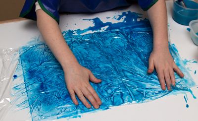 A child covering a sheet of paper with wet paint with a layer of cling film
